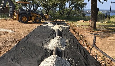 Wide video of a future Kickabout installation under construction, a flat surface enclosed by black liner and piles of drainstone in the foreground. In the back, a Hellas truck and a trailer stationed next to a two-story building.