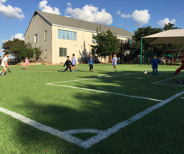 A class of young boys and girls happily running through a green synthetic turf Kickabout surface, as if just released for recess